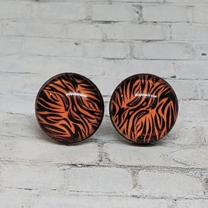 May include: A pair of round stud earrings with a tiger stripe design. The earrings feature a black and orange pattern. The earrings are set against a white brick background.