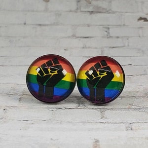 May include: A pair of round stud earrings. Each earring features a black fist graphic over a rainbow-colored background. The earrings are set against a white brick wall.