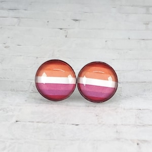 May include: Two round stud earrings featuring the lesbian pride flag design. The earrings display horizontal stripes of orange, pink, white, and magenta. The earrings are set against a white brick background.