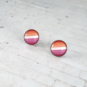 May include: A pair of round stud earrings featuring the lesbian pride flag design. The earrings have a glossy finish and a silver-colored metal border. The flag design includes stripes of orange, white, and pink.