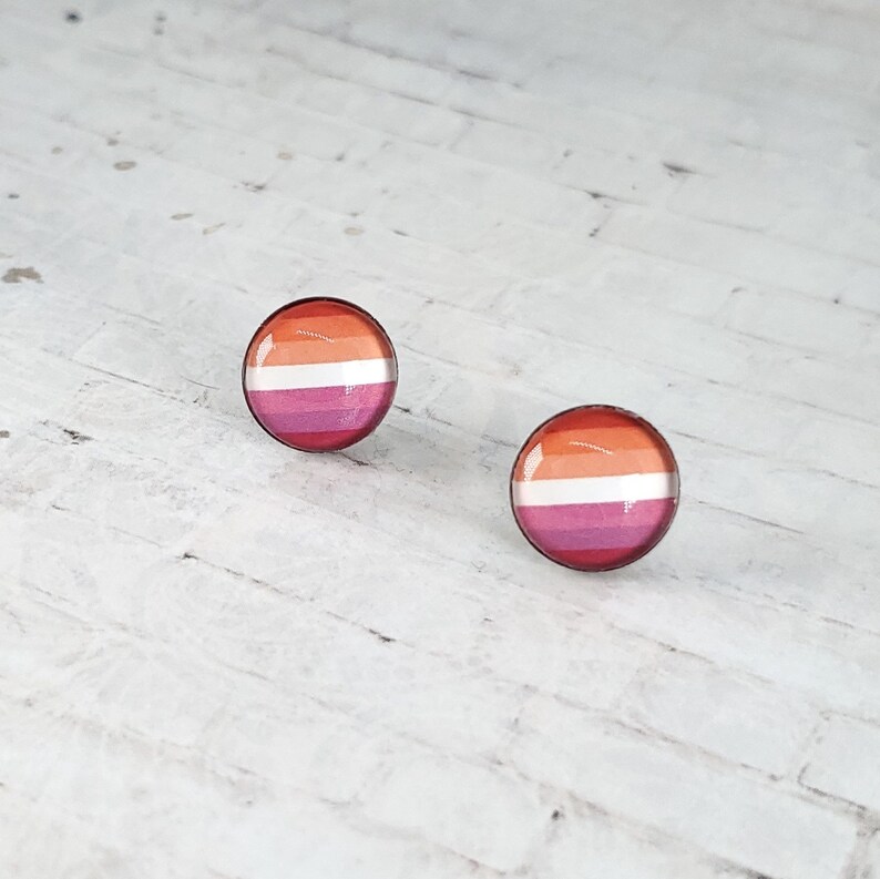 May include: A pair of round stud earrings featuring the lesbian pride flag design. The earrings have a glossy finish and a silver-colored metal border. The flag design includes stripes of orange, white, and pink.