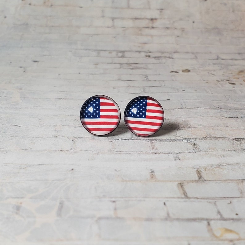 May include: A pair of round stud earrings featuring the American flag design. Each earring displays the red and white stripes and the blue field with white stars, set against a black border. The earrings are displayed on a white brick background.