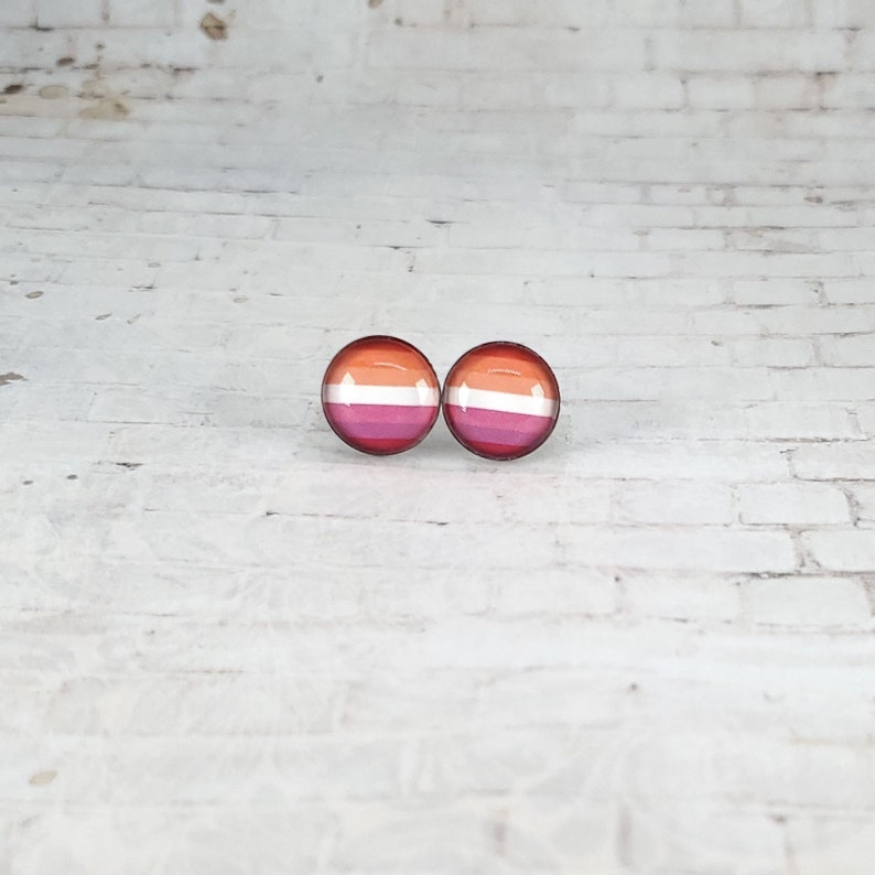 May include: Two round stud earrings featuring the lesbian pride flag design. The earrings display horizontal stripes of orange, pink, white, and magenta. The earrings are set against a white brick background.
