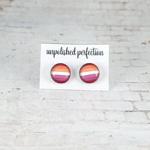 May include: A pair of round stud earrings featuring the lesbian pride flag design. The earrings display a gradient of colors, including orange, pink, and white stripes. The earrings are displayed on a white card with the text "unpolished perfection".