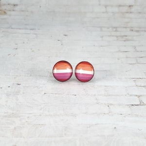 May include: Two round stud earrings with a lesbian pride flag design. The earrings feature a circular design with horizontal stripes in shades of orange, white, and pink. The earrings are set against a white brick background.