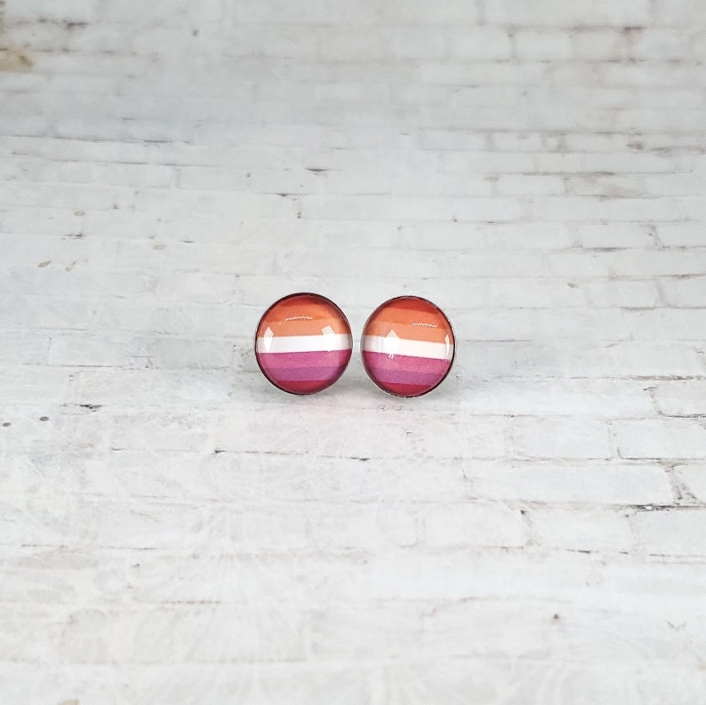 May include: Two round stud earrings with a lesbian pride flag design. The earrings feature a circular design with horizontal stripes in shades of orange, white, and pink. The earrings are set against a white brick background.
