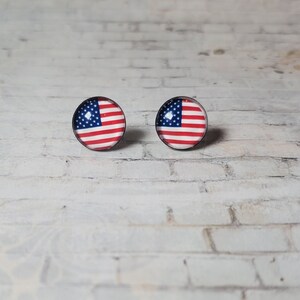 May include: A pair of round stud earrings featuring the American flag design. Each earring displays the stars and stripes of the flag within a circular, black-rimmed setting. The earrings are set against a white brick background.