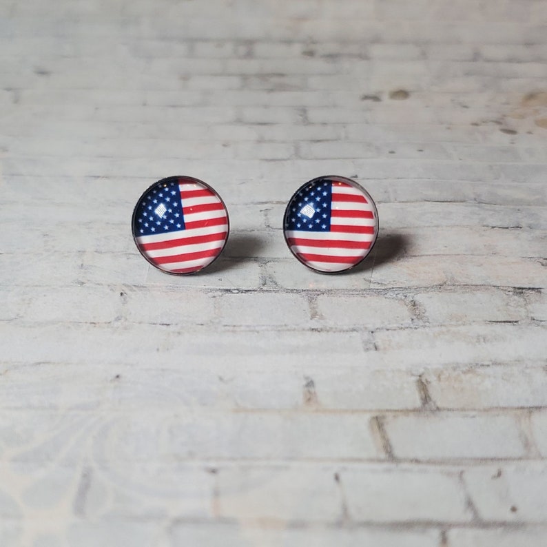May include: A pair of round stud earrings featuring the American flag design. Each earring displays the stars and stripes of the flag within a circular, black-rimmed setting. The earrings are set against a white brick background.