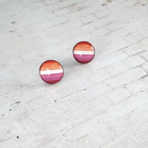 May include: Two round stud earrings with a glossy finish. Each earring features a horizontal striped design in shades of orange, white, pink, and magenta, representing the lesbian pride flag. The earrings are set against a white brick background.