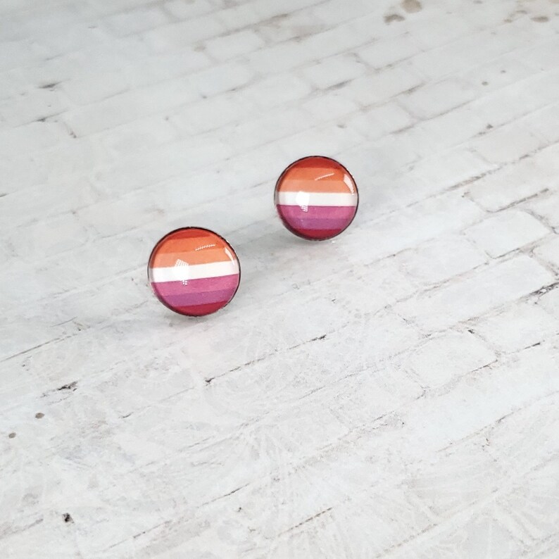 May include: Two round stud earrings with a glossy finish. Each earring features a horizontal striped design in shades of orange, white, pink, and magenta, representing the lesbian pride flag. The earrings are set against a white brick background.