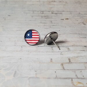 May include: A pair of round stud earrings featuring the American flag design. Each earring has a stainless steel post and a red, white, and blue flag graphic. The earrings are displayed on a white brick surface.