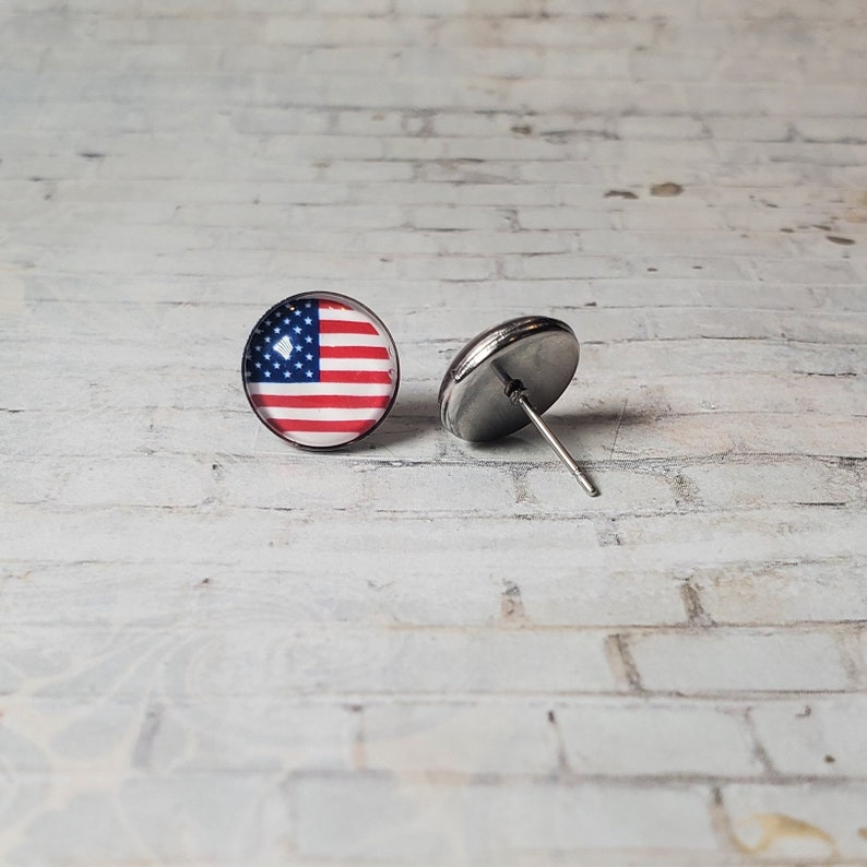 May include: A pair of round stud earrings featuring the American flag design. Each earring has a stainless steel post and a red, white, and blue flag graphic. The earrings are displayed on a white brick surface.