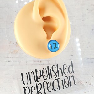 May include: A blue stud earring with the number 12 on a light skin-tone ear model. The earring is displayed on a clear acrylic stand with the words "Unpolished Perfection" and the text "*12mm size shown for sizing purposes only".