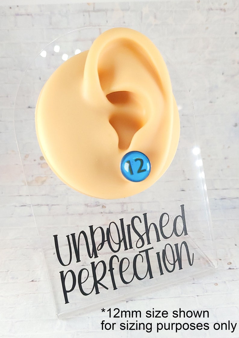 May include: A blue stud earring with the number 12 on a light skin-tone ear model. The earring is displayed on a clear acrylic stand with the words "Unpolished Perfection" and the text "*12mm size shown for sizing purposes only".