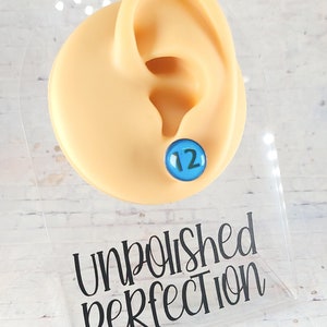 May include: A blue stud earring with the number 12 on a light skin-tone ear model. The earring is displayed on a clear acrylic stand with the words "Unpolished Perfection" and "*12mm size shown for sizing purposes only" in black text.