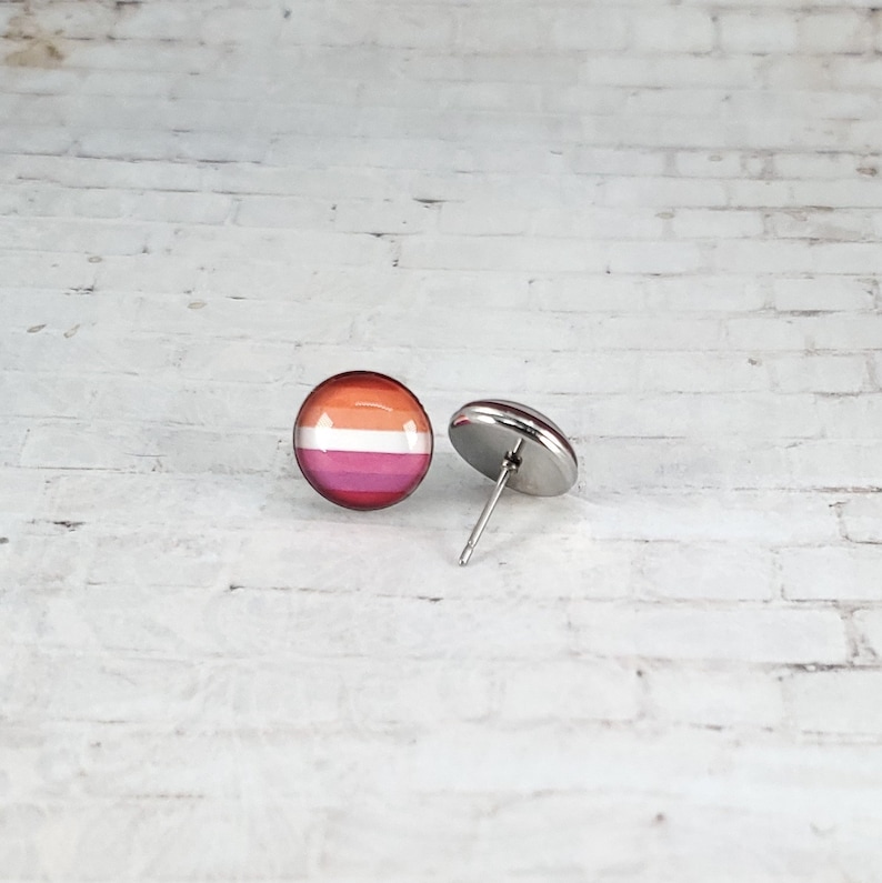 May include: A pair of stud earrings featuring the lesbian pride flag design. The earrings have a round, glossy surface with horizontal stripes in shades of orange, pink, and red. The posts and backs are silver-toned metal.