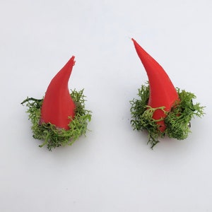 May include: Two red, conical horns, possibly for a costume, are surrounded by green moss. The horns are set against a white background, creating a striking contrast. The horns appear to be made of a solid material.