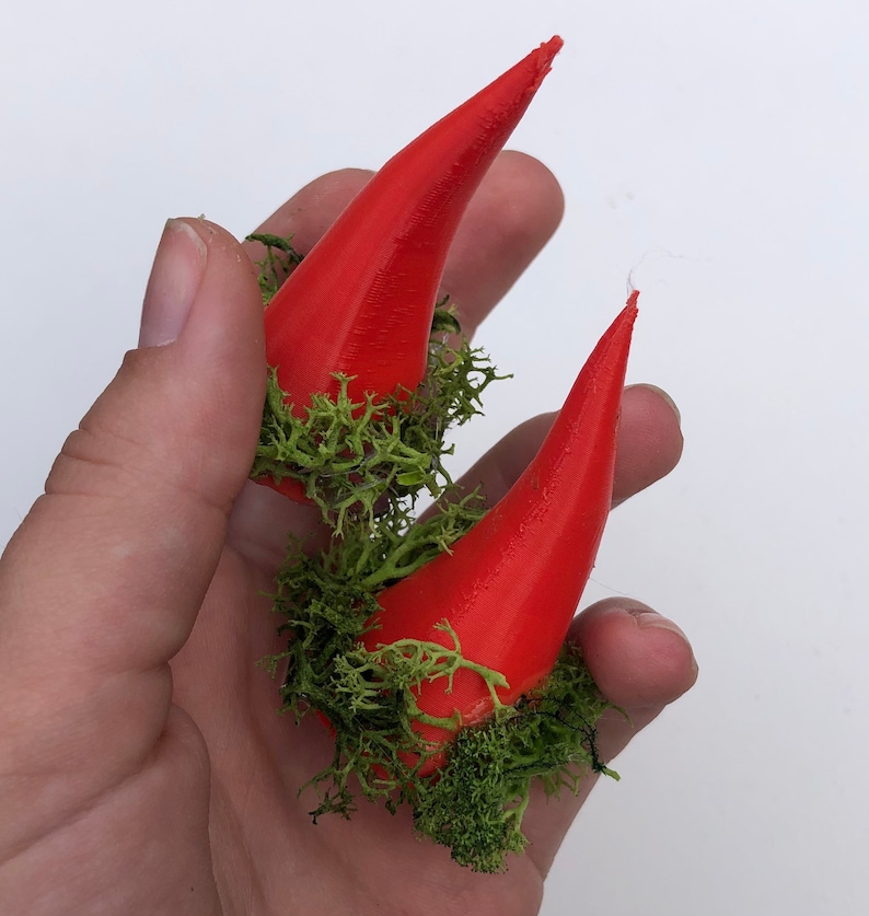May include: Two vivid red, conical horns with a textured surface, encircled by bright green moss. The horns are held in a hand against a white backdrop. These horns are likely for a costume or decorative use.