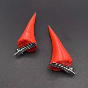 May include: A pair of red devil horn hair clips. The horns are a smooth, conical shape with a textured base. Each clip is made of silver-coloured metal. The horns are a vibrant red colour, perfect for a costume or themed event.