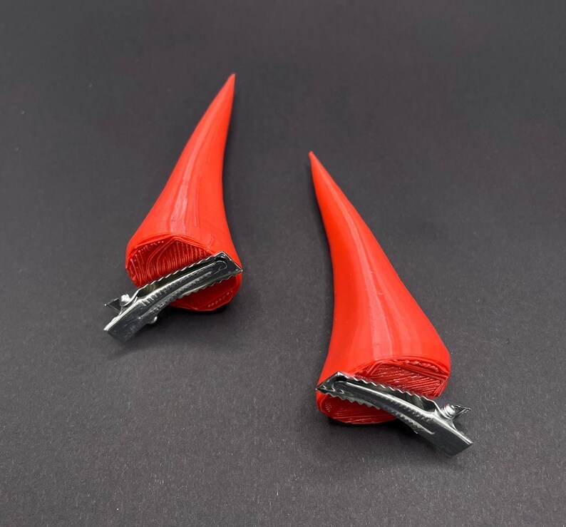 May include: A pair of red devil horn hair clips. The horns are a smooth, conical shape with a textured base. Each clip is made of silver-coloured metal. The horns are a vibrant red colour, perfect for a costume or themed event.