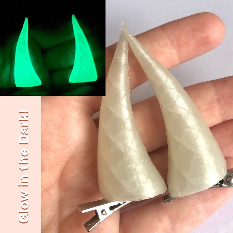 May include: Two off-white, textured unicorn horn hair clips. The horns are curved and have a pearlescent sheen. A smaller inset image shows the horns glowing bright green. Text on the left reads "Glow in the Dark!"