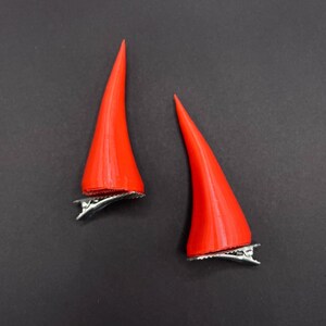 May include: Two bright red devil horn hair clips. Each horn is a smooth, conical shape with a silver metal clip attached to the base. The horns are set against a dark grey background.