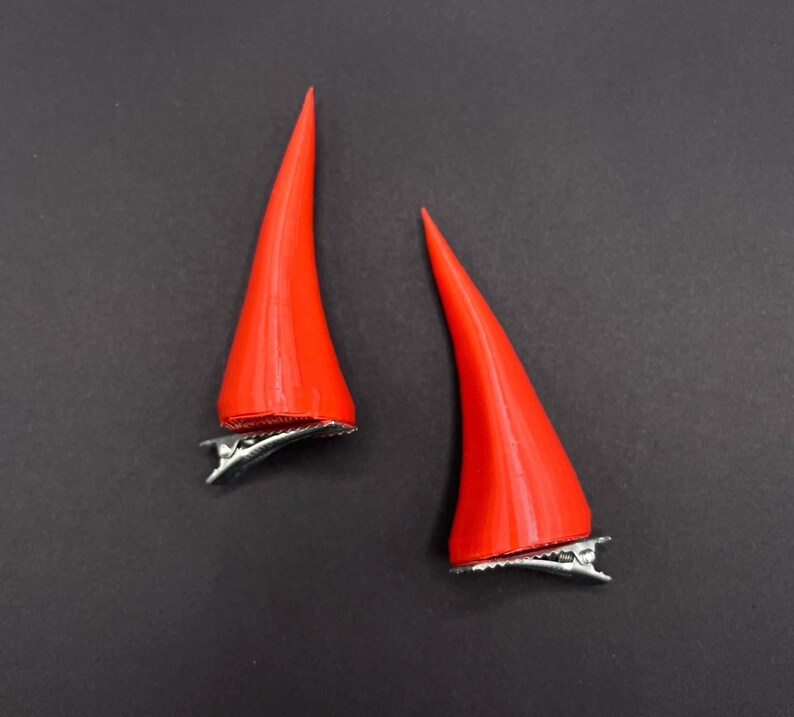 May include: Two bright red devil horn hair clips. Each horn is a smooth, conical shape with a silver metal clip attached to the base. The horns are set against a dark grey background.