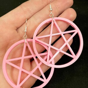 May include: A pair of pink earrings featuring a pentagram design. The earrings are circular with a five-pointed star inside. They have silver-colored ear wires.