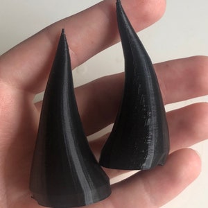 May include: Two black, cone-shaped horns with a slightly curved design. The horns are 3D-printed and have a textured surface. They are held in a hand, showcasing their size and shape. The horns are likely for cosplay or costume use.