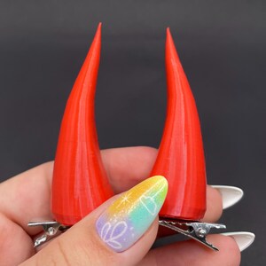 May include: Two vivid red devil horns, each secured with a silver metal alligator clip. The horns are a striking shade of red and have a textured finish. These clips are designed for easy attachment to hair, providing a fun accessory.