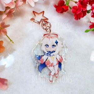 Op de afbeelding: Een transparante acryl sleutelhanger met een anime-achtig personage met wit haar, een rood-witte outfit en blauwe vleugels. Het personage staat op een blauw-witte achtergrond met sterren. De sleutelhanger heeft een gouden ster-bedel bevestigd aan een metalen ring.