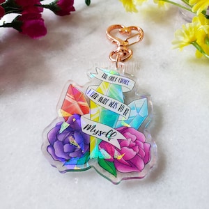 May include: A clear iridescent keychain with a pink, purple, and blue flower design. The keychain features a quote that reads "The only choice I ever made was to be myself."