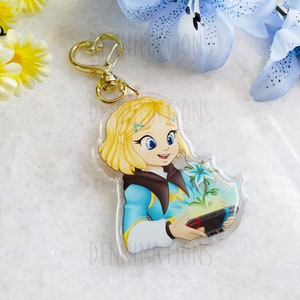 Puede incluir: Un llavero de acrílico transparente con una ilustración de dibujos animados de una joven con cabello rubio, vestida con un atuendo azul y marrón, sosteniendo un mando de videojuegos y una flor blanca. El fondo es un degradado de azul claro a blanco.