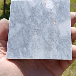 May include: A square, grey marble tile with a subtle, veined pattern. The tile is held in a hand, showcasing its smooth surface and natural colour variations. The marble has a polished finish.