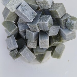 May include: A collection of small, rectangular, and cube-shaped stones in varying shades of gray and green. The stones are piled together in a white container, creating a textured and natural appearance. These stones could be used for crafts or decorative purposes.