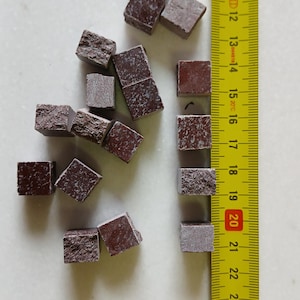 May include: A collection of small, reddish-brown, square-shaped stone tiles scattered on a white surface. A yellow ruler with black markings is positioned next to the tiles, showing measurements in inches.