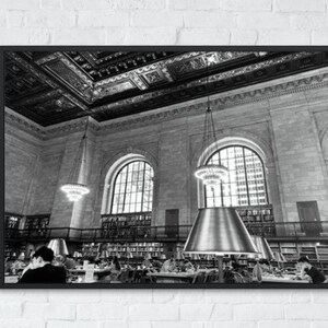 Puede incluir: Fotografía en blanco y negro del interior de una biblioteca con techos altos, detalles ornamentados y grandes ventanales. Personas sentadas en mesas leyendo libros bajo grandes pantallas de lámparas plateadas.