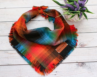 May include: A plaid dog bandana with frayed edges in shades of orange, blue, green, and black. The bandana has a brown leather tag with the text "Maxie's Designs" on it.