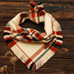 May include: A triangular pet bandana featuring a plaid pattern in cream, red, and black. The fabric has a fringed edge and a gold-tone metal snap closure at the top. A small brown leather tag with the text Maxxie's Designs is sewn onto the fabric.
