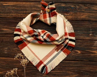 May include: A triangular pet bandana featuring a plaid pattern in cream, red, and black. The fabric has a fringed edge and a gold-tone metal snap closure at the top. A small brown leather tag with the text Maxxie's Designs is sewn onto the fabric.
