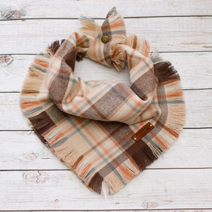May include: A brown, beige, and orange plaid dog bandana with frayed edges. The bandana has a brown leather tag with the text "Maxxie &  Designs" on it.
