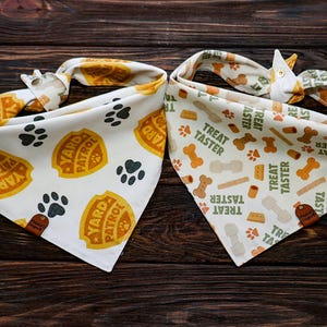 May include: Two dog bandanas on a wooden surface. One is white with orange "Yard Patrol" badges and paw prints. The other is white with dog bones, paw prints, and "Treat Taster" text. Both have a patterned tie.