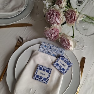 Puede incluir: Elegante mesa con platos blancos, cubiertos dorados y una servilleta de lino blanca. Tarjetas de nombre decorativas azules y blancas con los nombres "Nour" y "Kushal" colocadas sobre la servilleta. Flores rosas y blancas y copas de vino completan el arreglo.