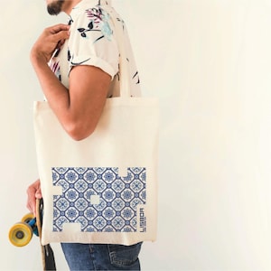 Op de afbeelding: Een witte canvas tote bag met een blauw en wit patroon met het woord "LISBOA" erop gedrukt. De tas wordt gedragen door iemand die een bloemenoverhemd en een blauwe jeans draagt.