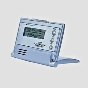 May include: A silver-colored digital clock radio with a fold-out stand. The clock face displays the time, date, and temperature. The radio has buttons for volume, mode, and set.