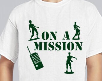 Puede incluir: Una camiseta blanca de manga corta con gráficos en color verde oscuro. El diseño incluye el texto ON A MISSION en fuente negrita, rodeado por siluetas de tres figuras en poses de acción y un dispositivo de radio walkie-talkie portátil.