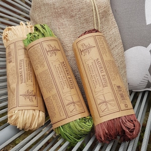 May include: Three spools of natural raffia ribbon in brown, green, and natural colors. The spools are wrapped in brown paper with the text "Natural Raffia" printed on them. The spools are sitting on a gray chair with a burlap bag behind them.