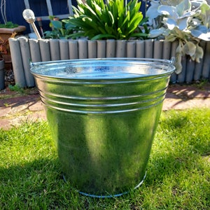 May include: A silver galvanised metal bucket with a rolled rim and two handles. The bucket is sitting on a bed of green grass.