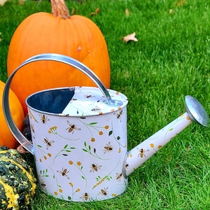 May include: A white metal watering can with a bee and floral pattern. The can has a silver handle and spout. The background includes pumpkins and green grass, suggesting a fall or garden theme.