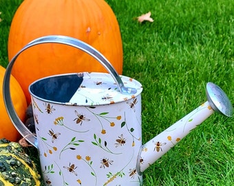 May include: A white metal watering can decorated with bees and floral motifs. It features a silver handle and spout. The image includes pumpkins and green grass, evoking a garden or autumn setting.